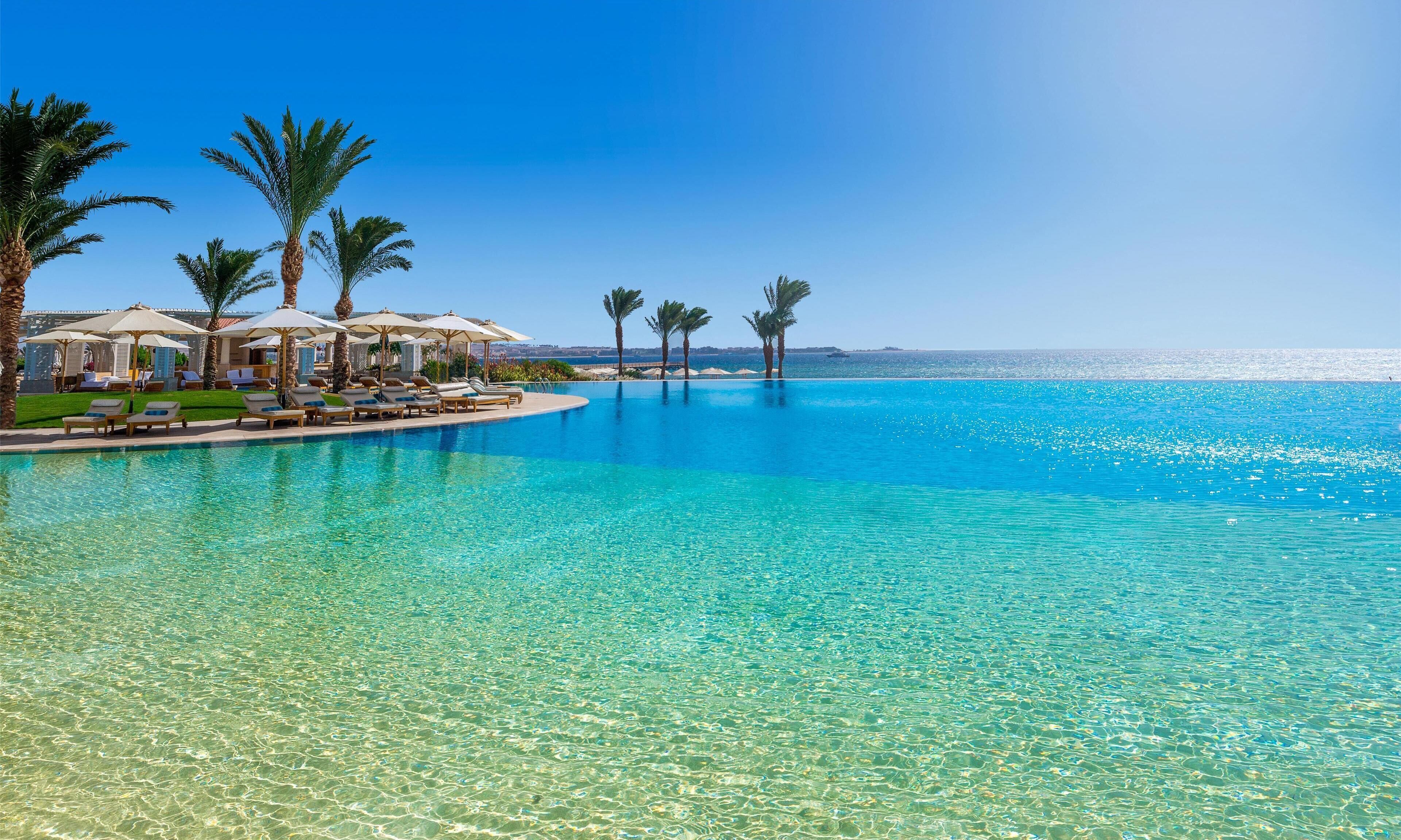 Una piscina cristallina blu con lettini e ombrelloni, palme e vista sul mare.