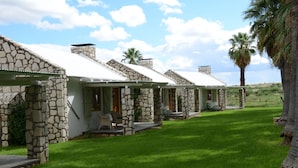 A row of stone-roofed cottages with a grassy lawn and palm trees. - Kalahari Farmhouse (Strampriet)