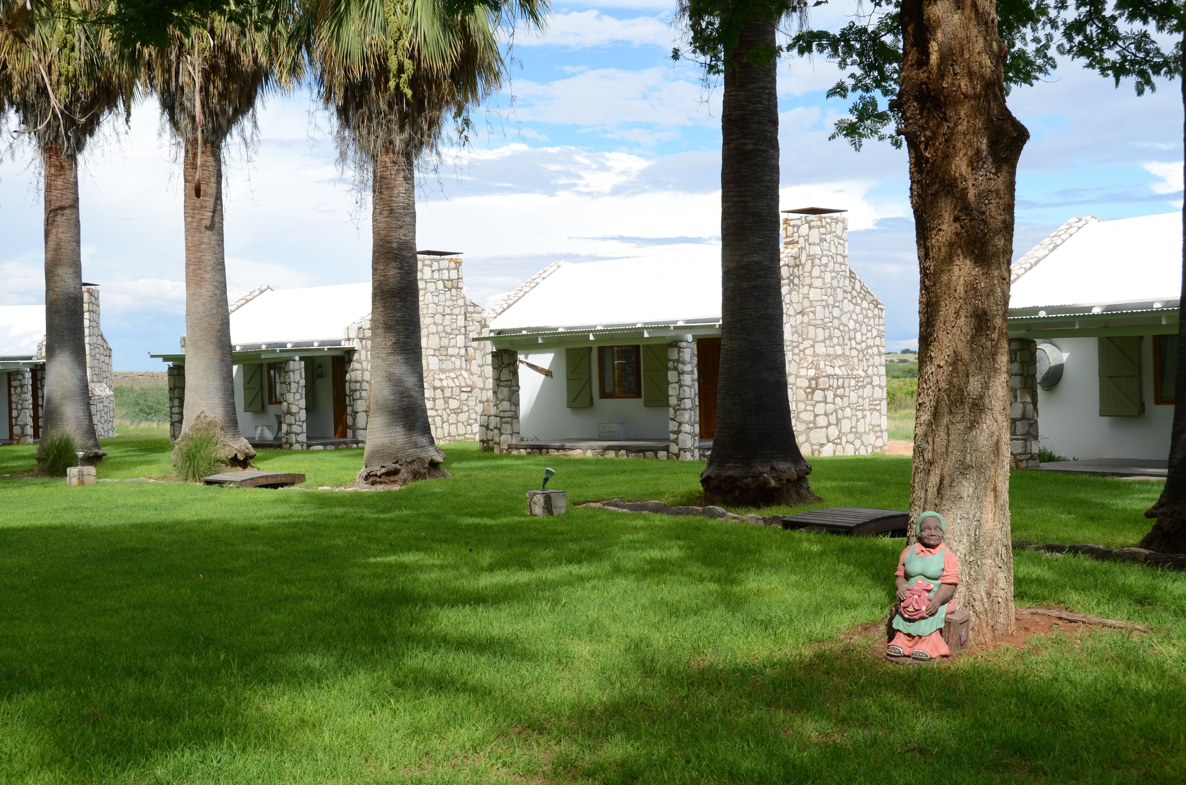 A row of stone cottages with green shutters, surrounded by lush grass and tall trees.