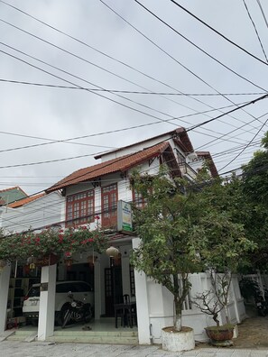 Il s’agit d’un bâtiment traditionnel, doté d’un toit en tuiles et d’un balcon. Une moto est garée devant, et un pot de fleurs se trouve à proximité.