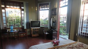 A hotel room with a bed, a TV, a desk, and a chair. There are windows with a view of greenery outside. - Homestay Countryside (Da Nang)