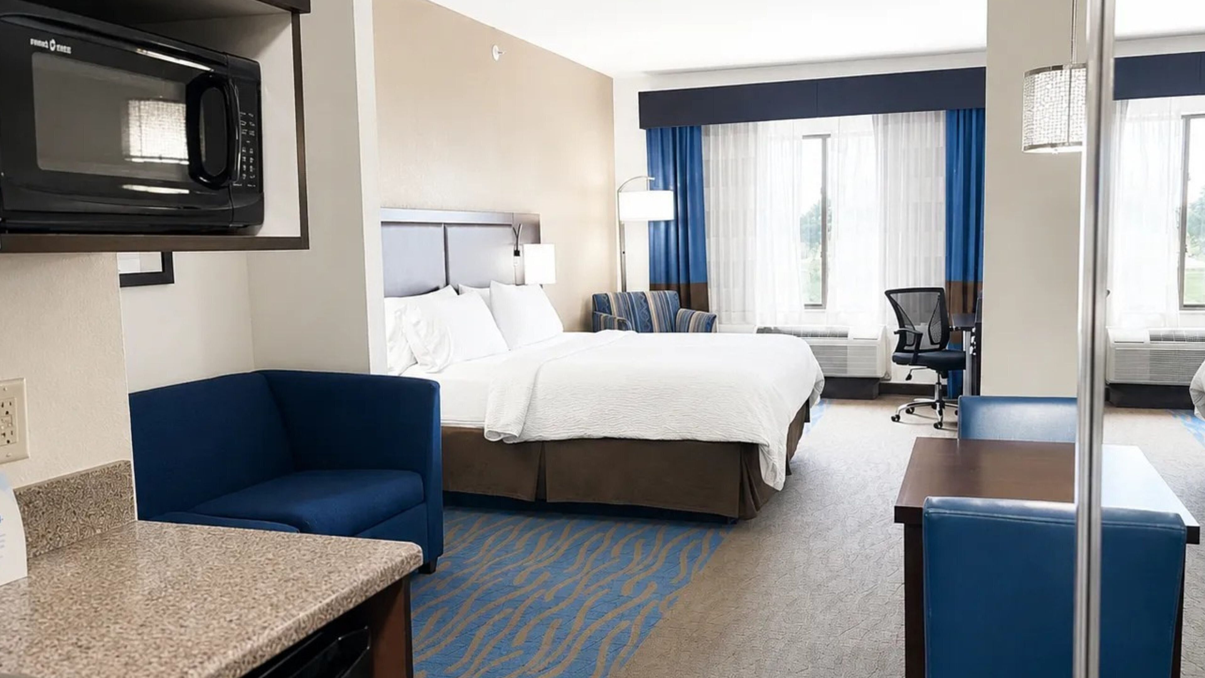A hotel room with a bed, a desk, a chair, a microwave, and a window with curtains.