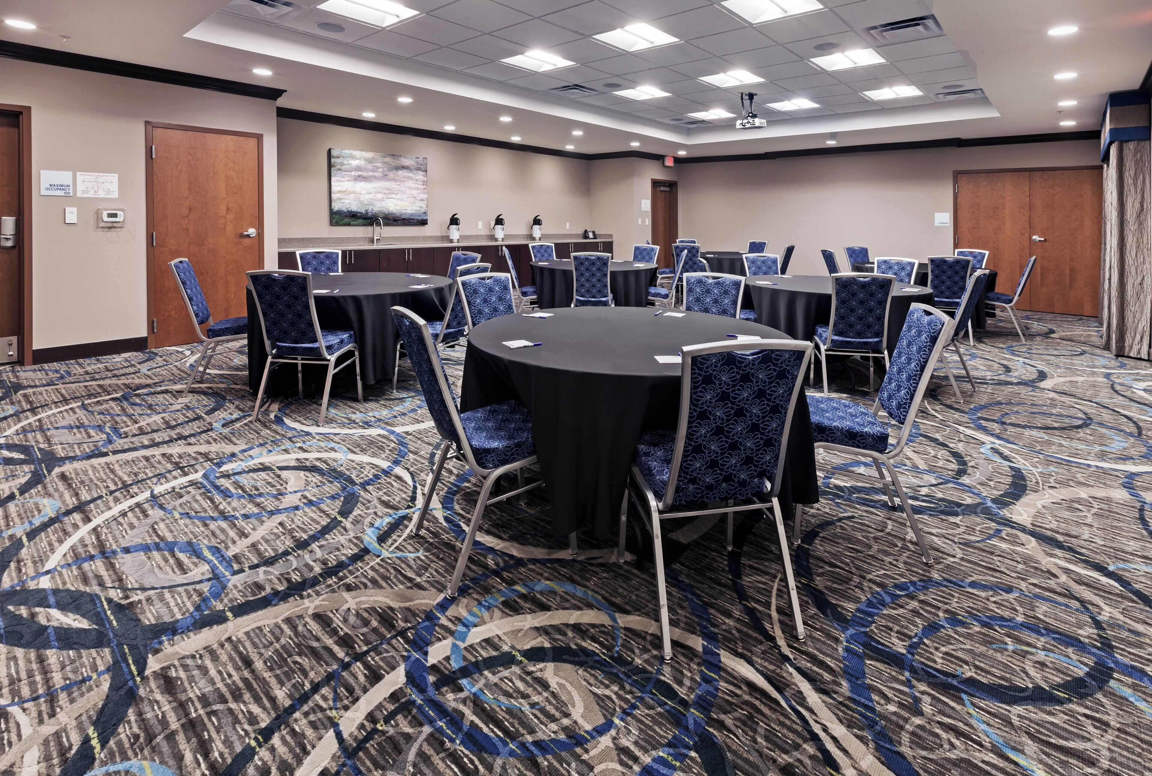 Une salle de conférence avec des tables rondes et des chaises à motifs bleus, un sol recouvert de moquette et un tableau accroché au mur.