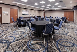 Un salón de conferencias con mesas redondas y sillas azules con estampado, suelo alfombrado y un cuadro en la pared.
