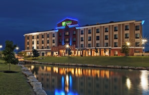 A Holiday Inn Express & Suites hotel with a reflection in a body of water, surrounded by a well-maintained lawn and a stone border. - Holiday Inn Express & Suites Glenpool-Tulsa South by IHG (Glenpool)