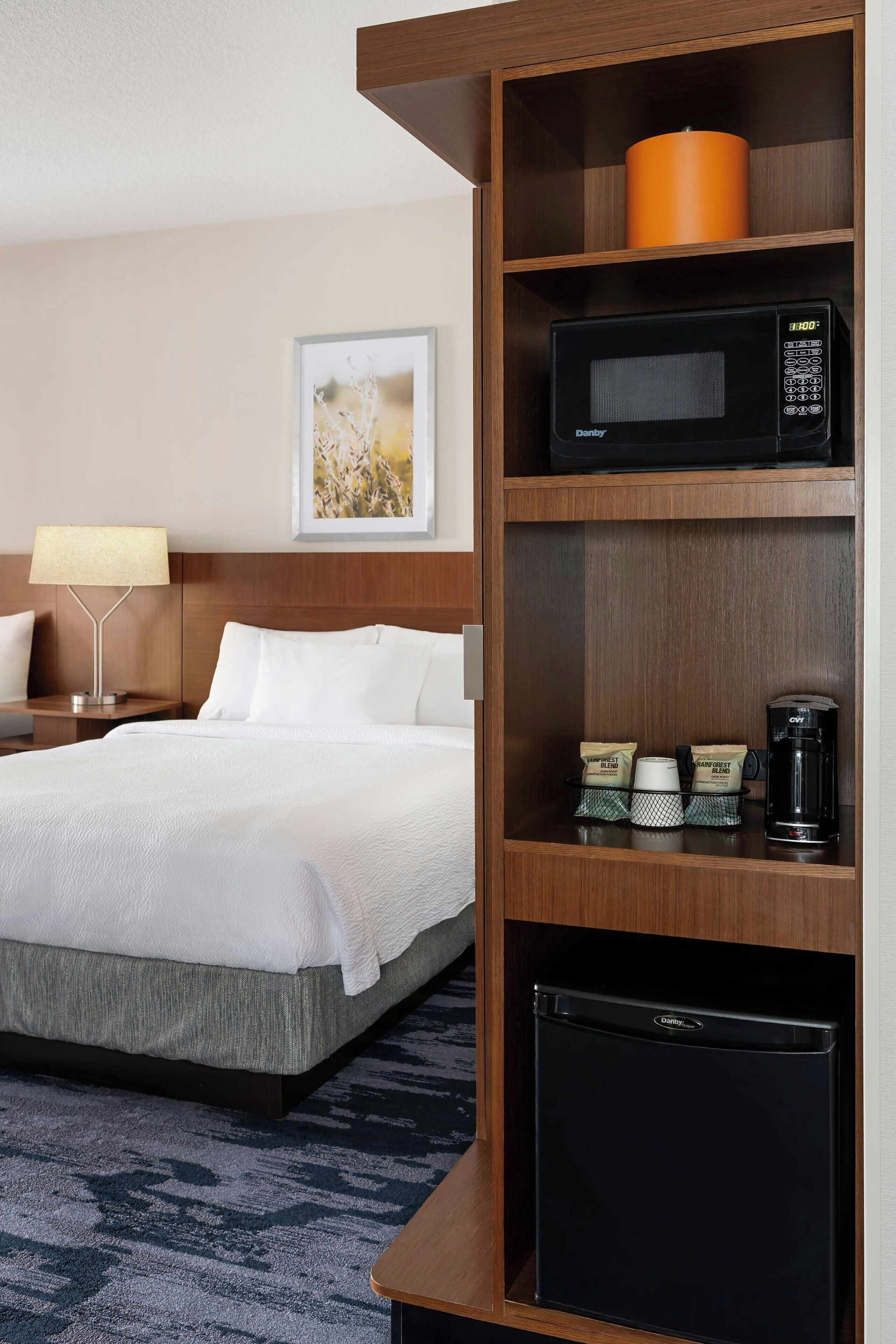 A hotel room with a bed, a nightstand with a lamp, a microwave, a coffee maker, and a small refrigerator.