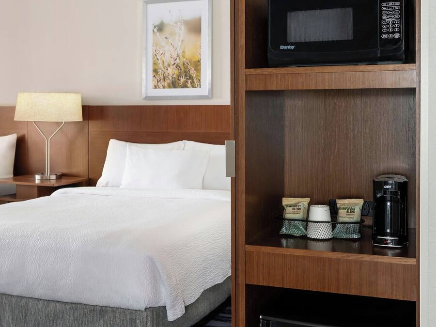 A hotel room with a bed, a nightstand with a lamp, a microwave, a coffee maker, and a small refrigerator.