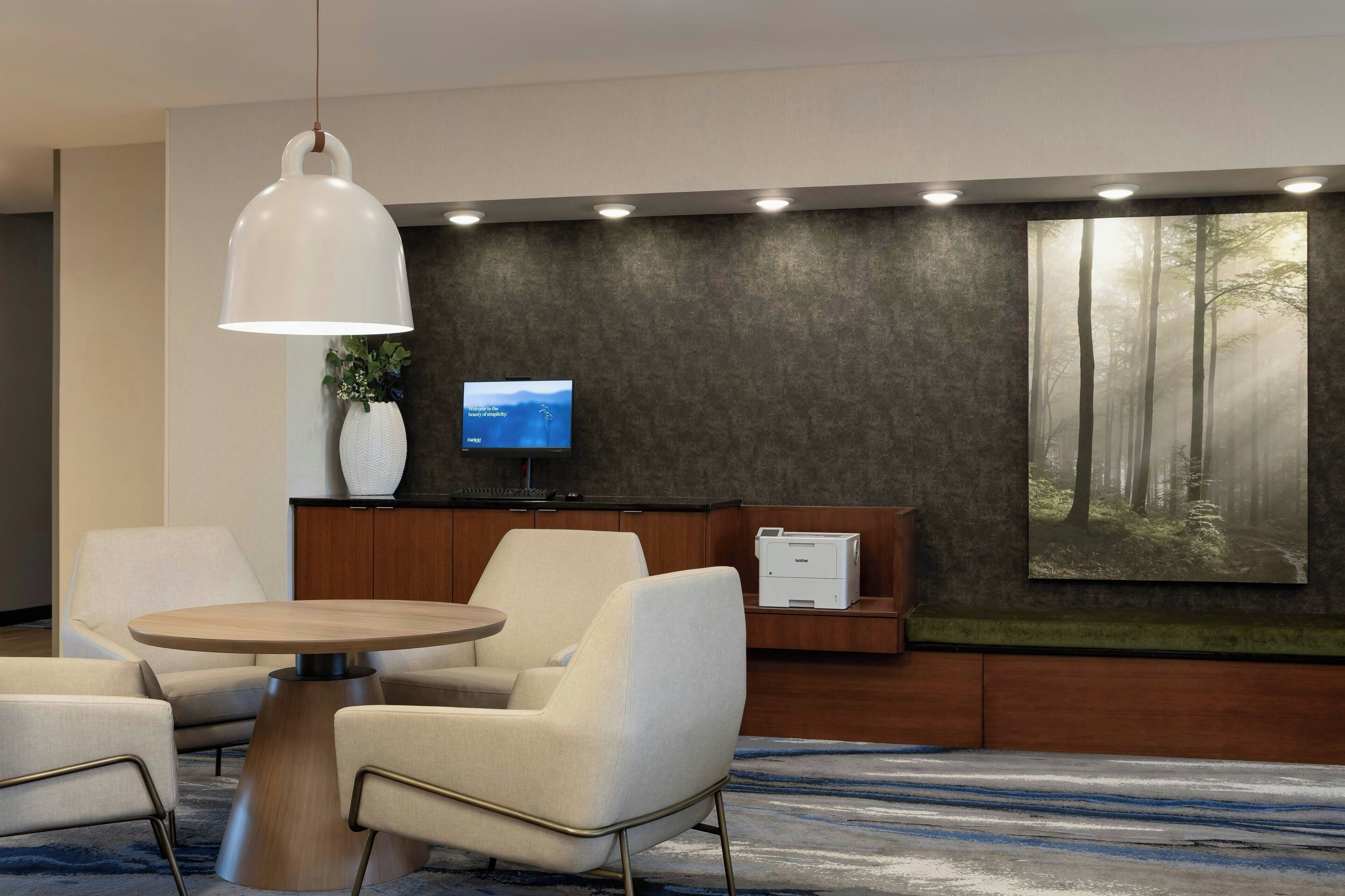 A modern lobby with a round wooden table, two white armchairs, a white pendant light, a flat-screen TV, a large forest mural, and a printer on a wooden cabinet.