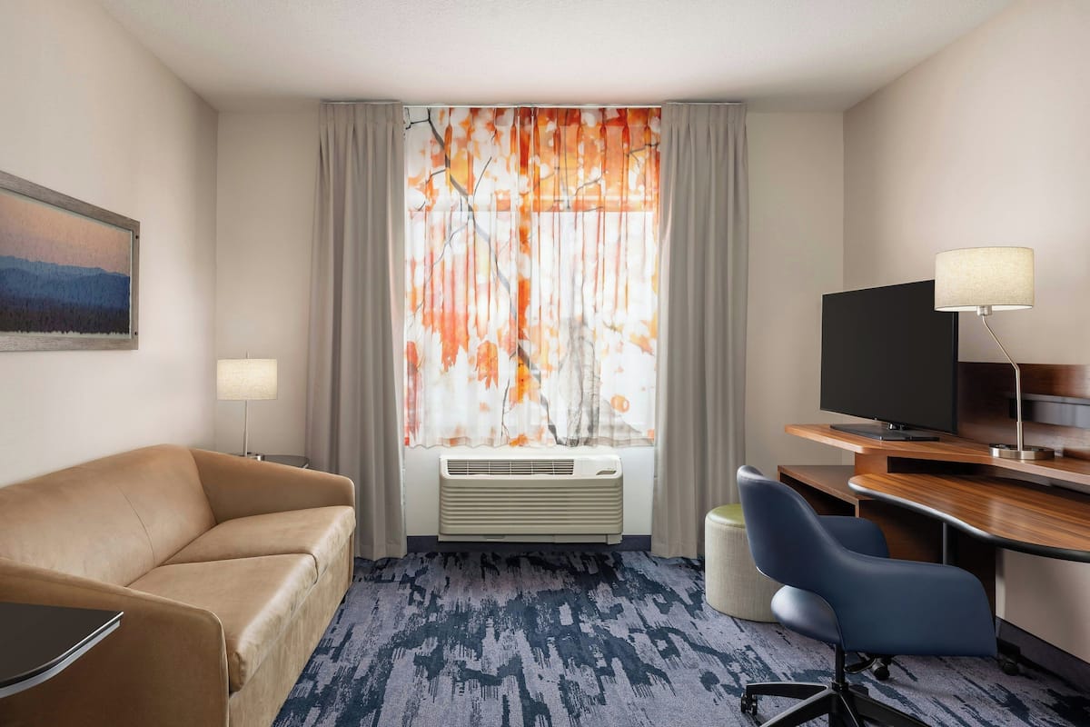Une chambre d’hôtel avec un canapé, un bureau et une chaise, une télévision et une fenêtre avec des rideaux.