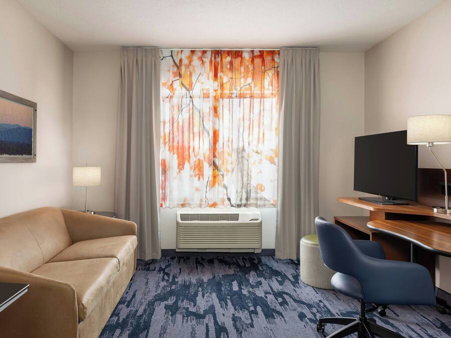 Une chambre d’hôtel avec un canapé, un bureau et une chaise, une télévision et une fenêtre avec des rideaux.
