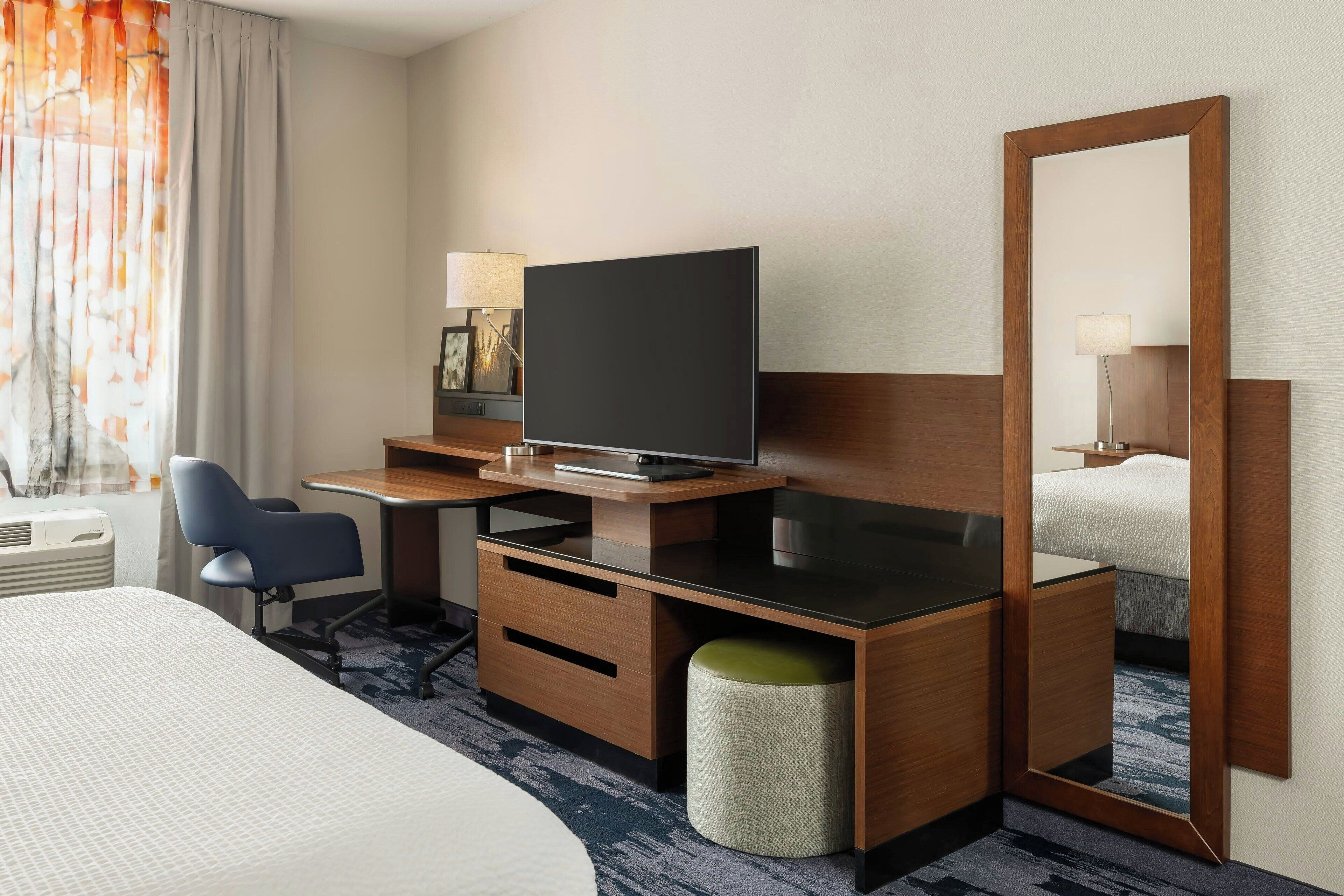A hotel room with a bed, a desk with a television, a chair, a mirror, and a lamp.