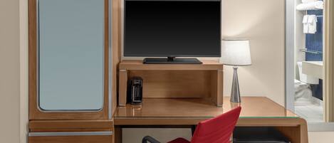 A hotel room with a wooden desk, a flat-screen TV, a red chair, a small refrigerator, and a coffee maker.