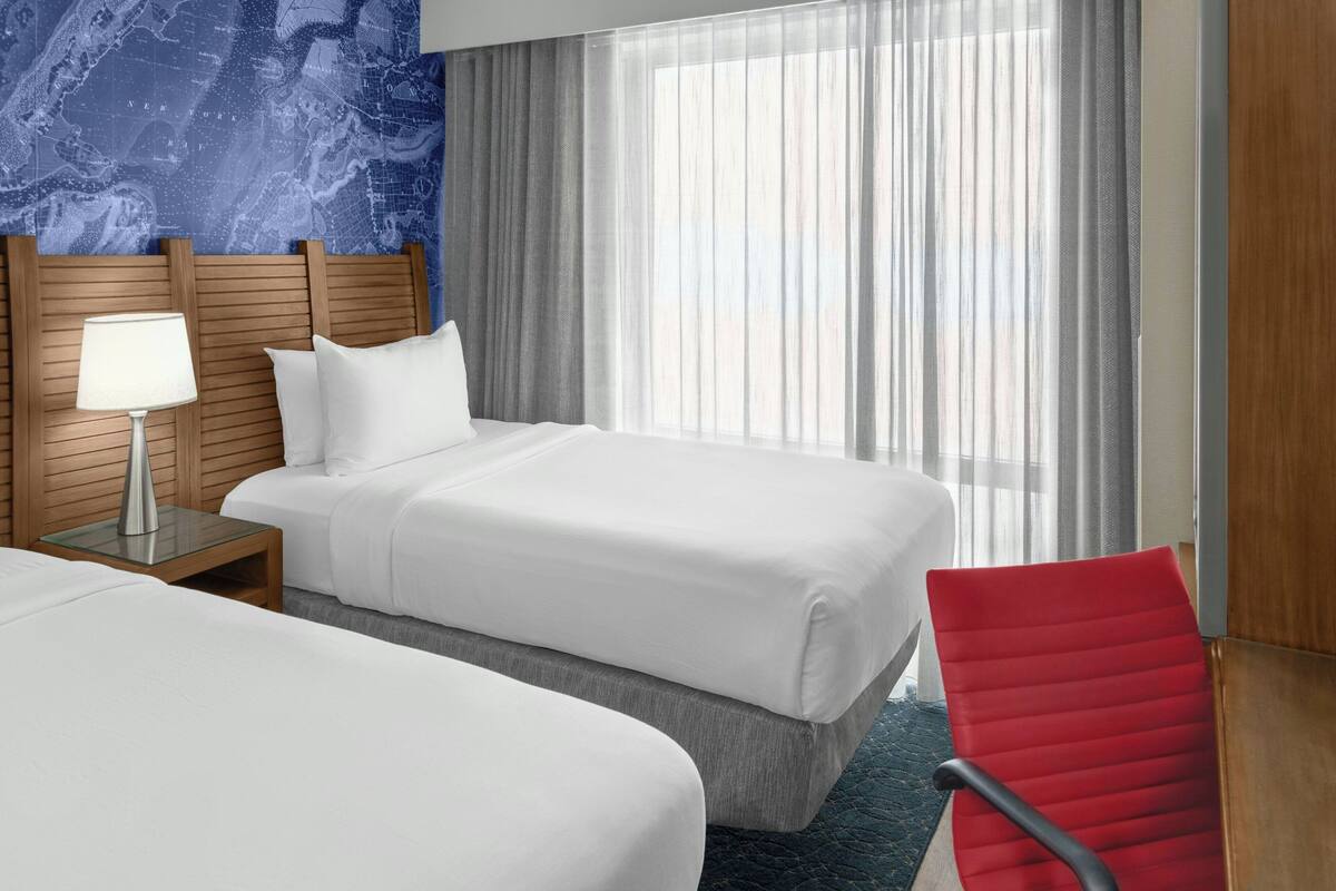 Een hotelkamer met twee bedden, een houten hoofdbord, een nachtkastje met een lamp, een rode stoel en een groot raam met dunne gordijnen.