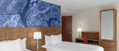 A hotel room with two beds, a large map mural, a wooden headboard, a desk with a lamp, a mirror, and a red chair.