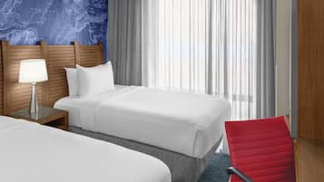 A hotel room with two beds, a wooden headboard, a nightstand with a lamp, a red chair, and a large window with sheer curtains.