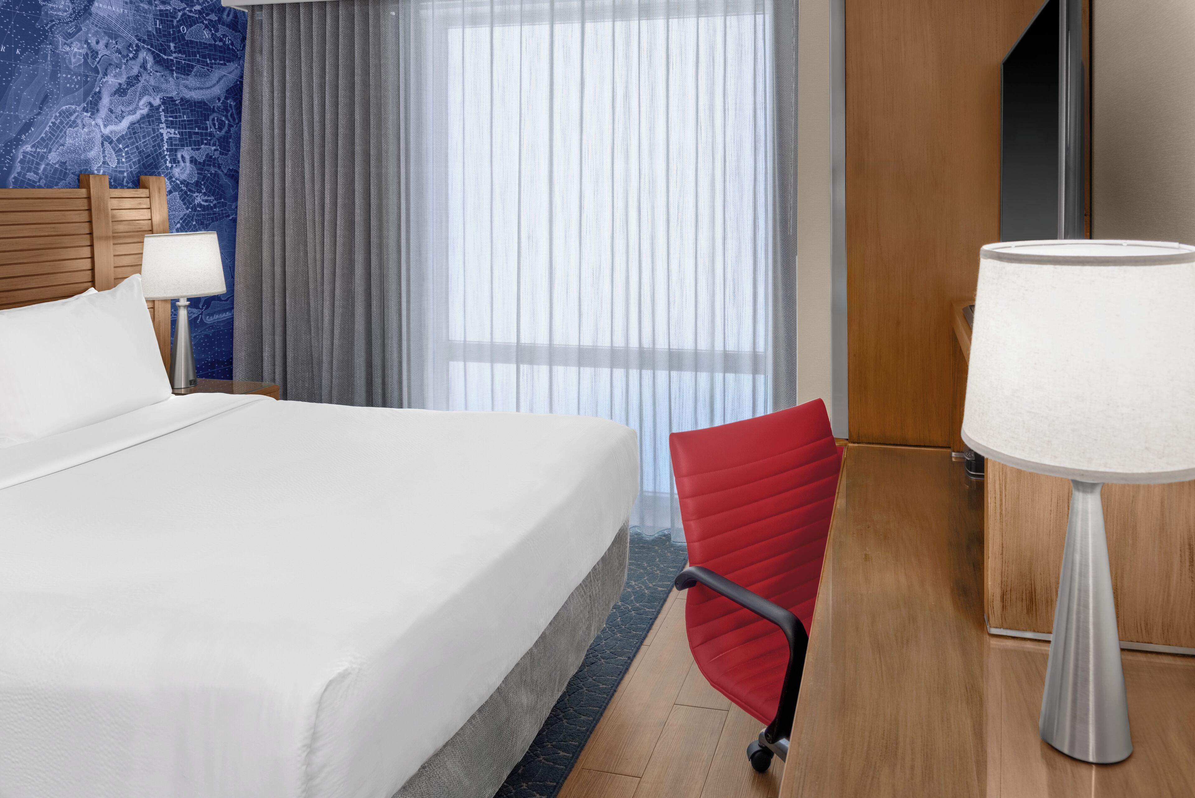 Una habitación de hotel con una cama grande, una silla de escritorio roja, un escritorio con una lámpara y una ventana con cortinas translúcidas.