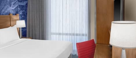 Una habitación de hotel con una cama grande, una silla de escritorio roja, un escritorio con una lámpara y una ventana con cortinas translúcidas.