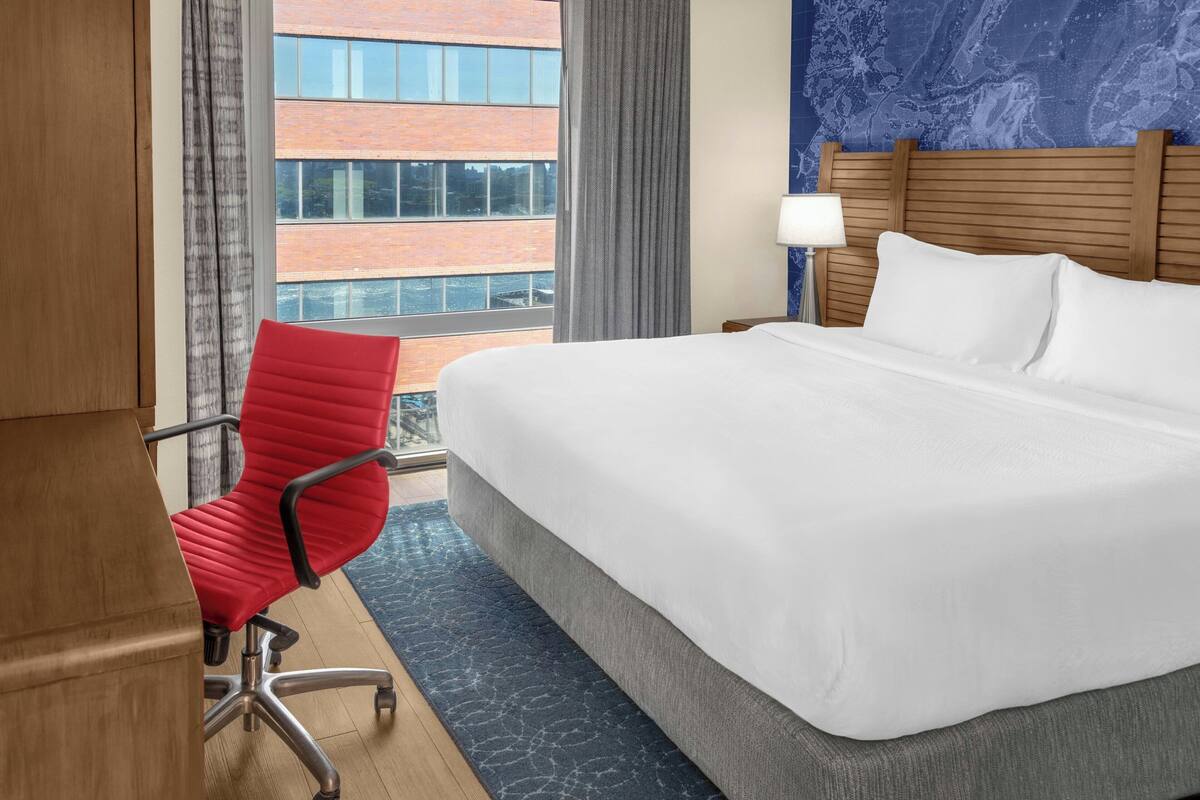 Hotelkamer met een groot bed, een rode bureaustoel, een nachtkastje met een lamp en uitzicht op een bakstenen gebouw.