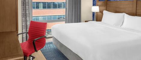 Quarto de hotel com uma cama grande, uma cadeira de escritório vermelha, uma mesa de cabeceira com abajur e vista para um edifício de tijolos pela janela.