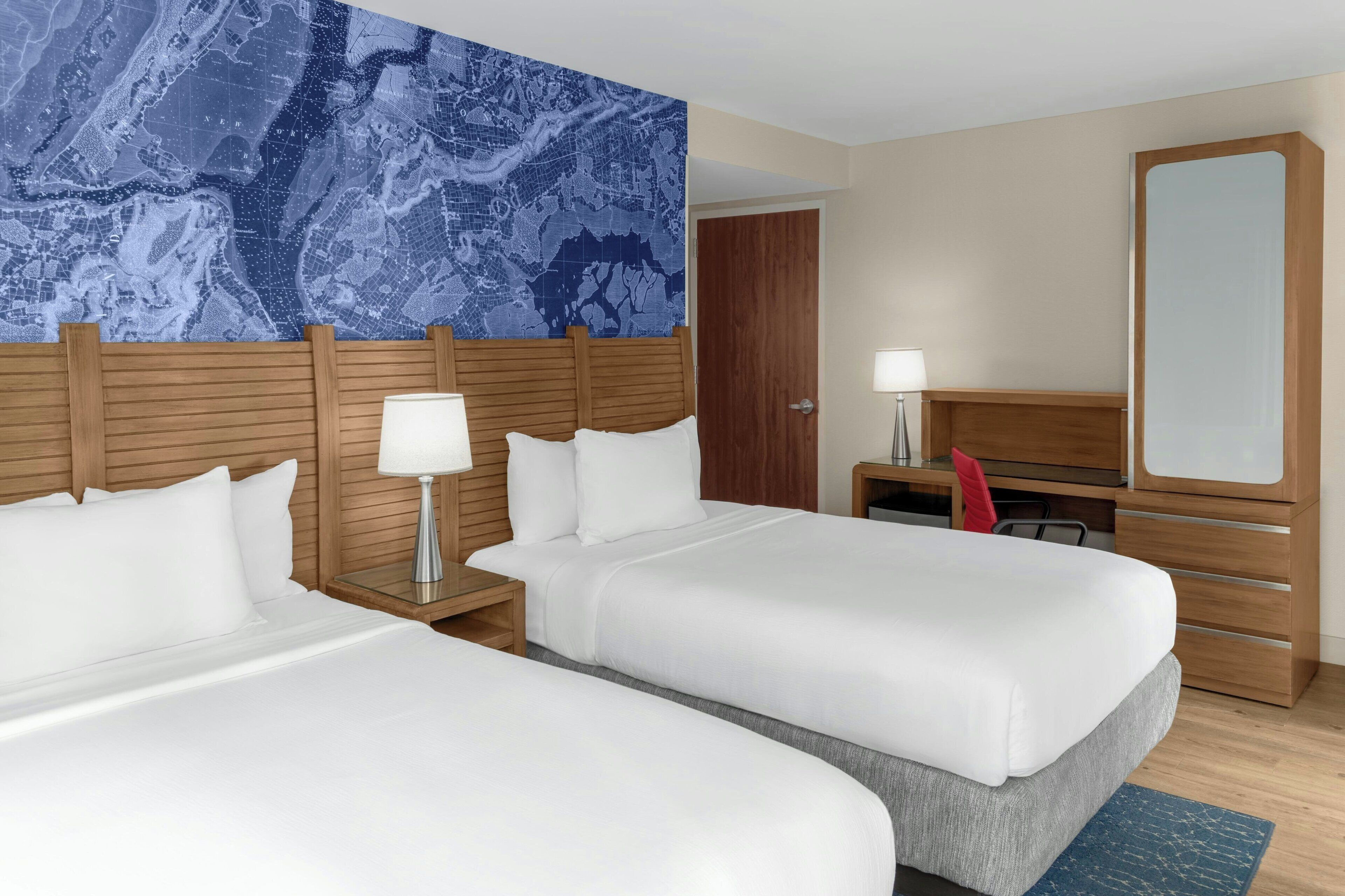 A hotel room with two beds, a large wall map, a wooden headboard, a desk with a lamp, a mirror, and a red chair.