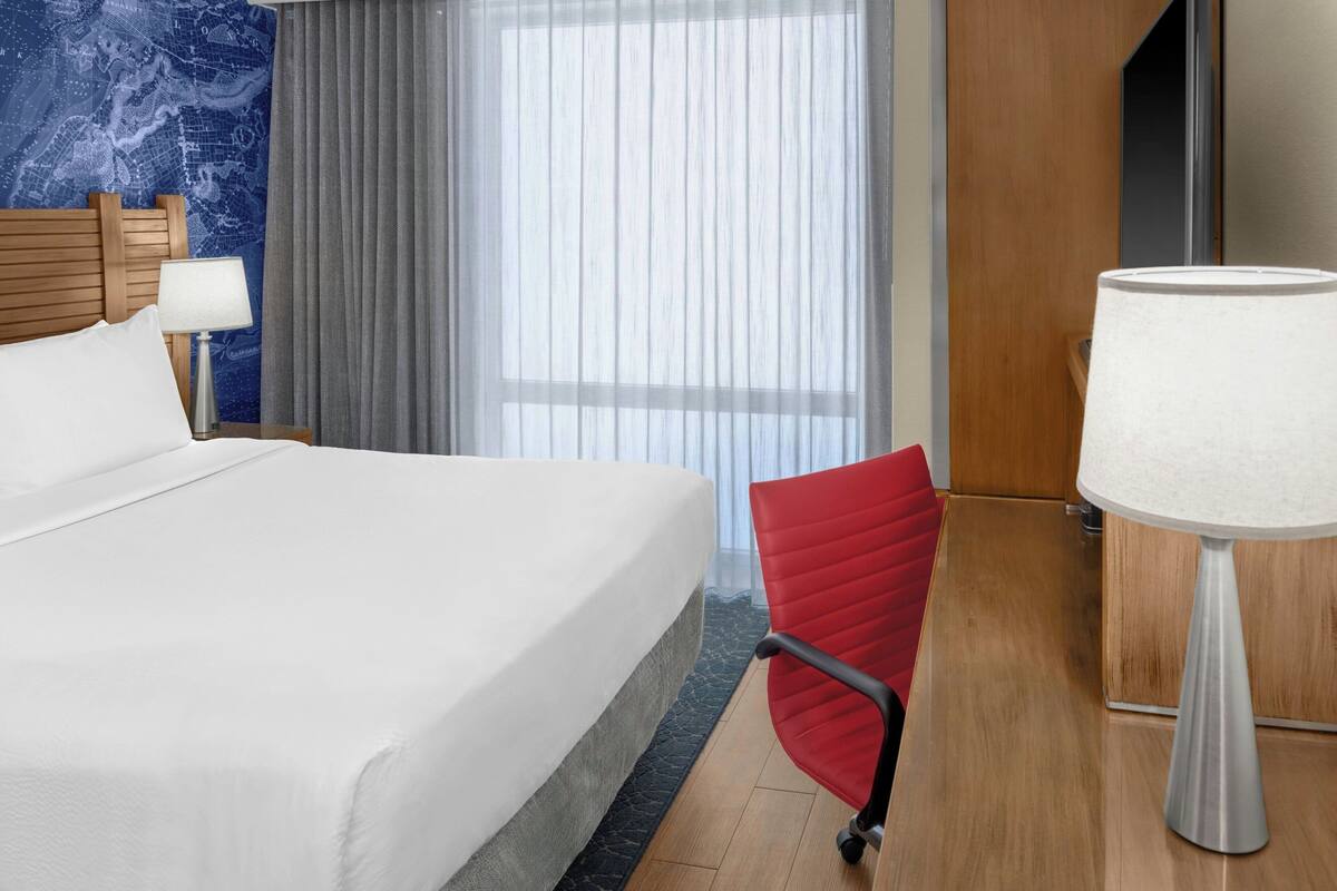 Hotelkamer met een groot bed, een rode stoel, een bureau met lamp en een raam met dunne gordijnen.