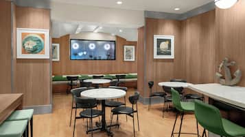 Een restaurant met ronde tafels en groene stoelen, houten wanden en kunstwerken in lijsten.