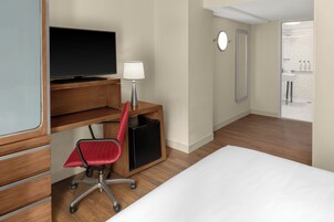 A hotel room with a bed, a desk with a computer and a chair, a TV, a lamp, and a bathroom visible through an open door.