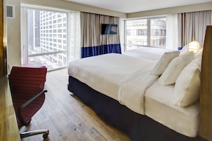 Quarto de hotel com uma cama grande, uma televisão na parede, uma cadeira vermelha e vista da cidade por grandes janelas.