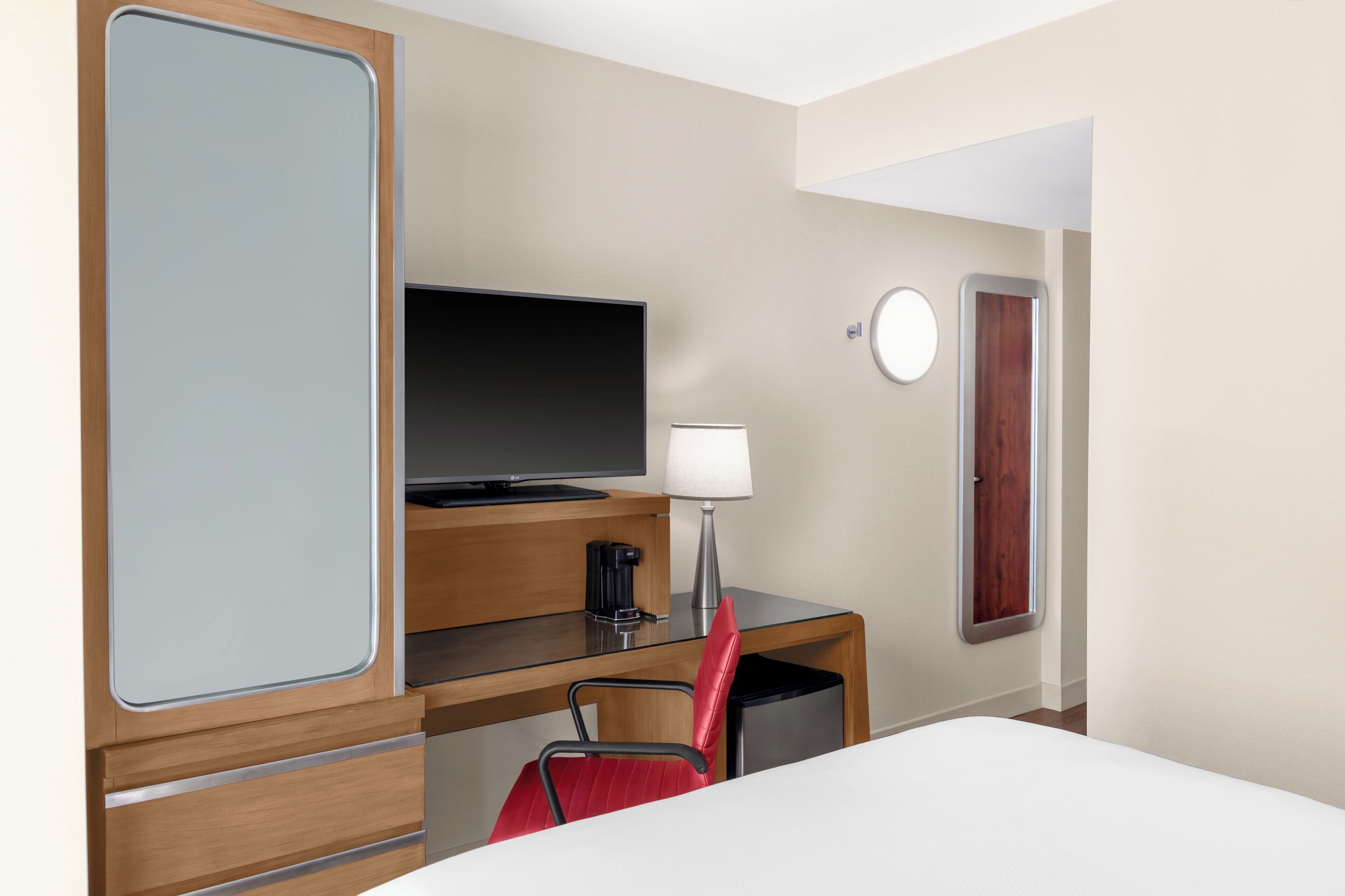 A hotel room with a bed, a desk with a TV, a lamp, a red chair, and a door with a window.