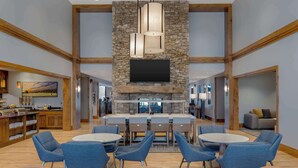 A spacious indoor area with a stone fireplace, a flat-screen TV mounted above it, and a dining area with blue chairs and round tables. - Homewood Suites By Hilton Billings, MT (Billings)