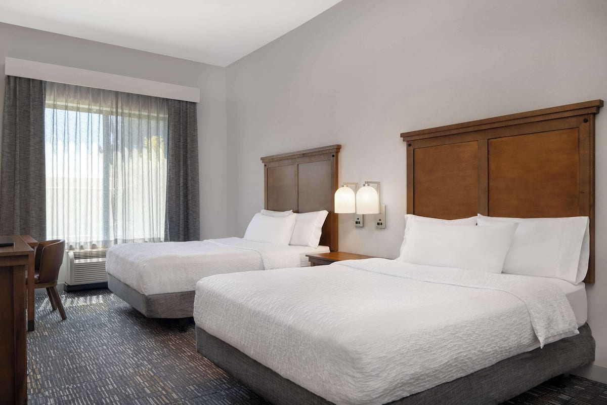 Een hotelkamer met twee bedden, een bureau en een stoel. Er zijn twee wandlampen en een raam met gordijnen.