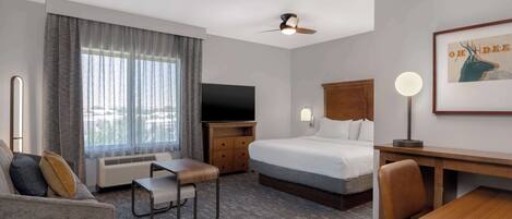 A hotel room with a bed, a desk, a chair, a television, a ceiling fan, and a large window with curtains.