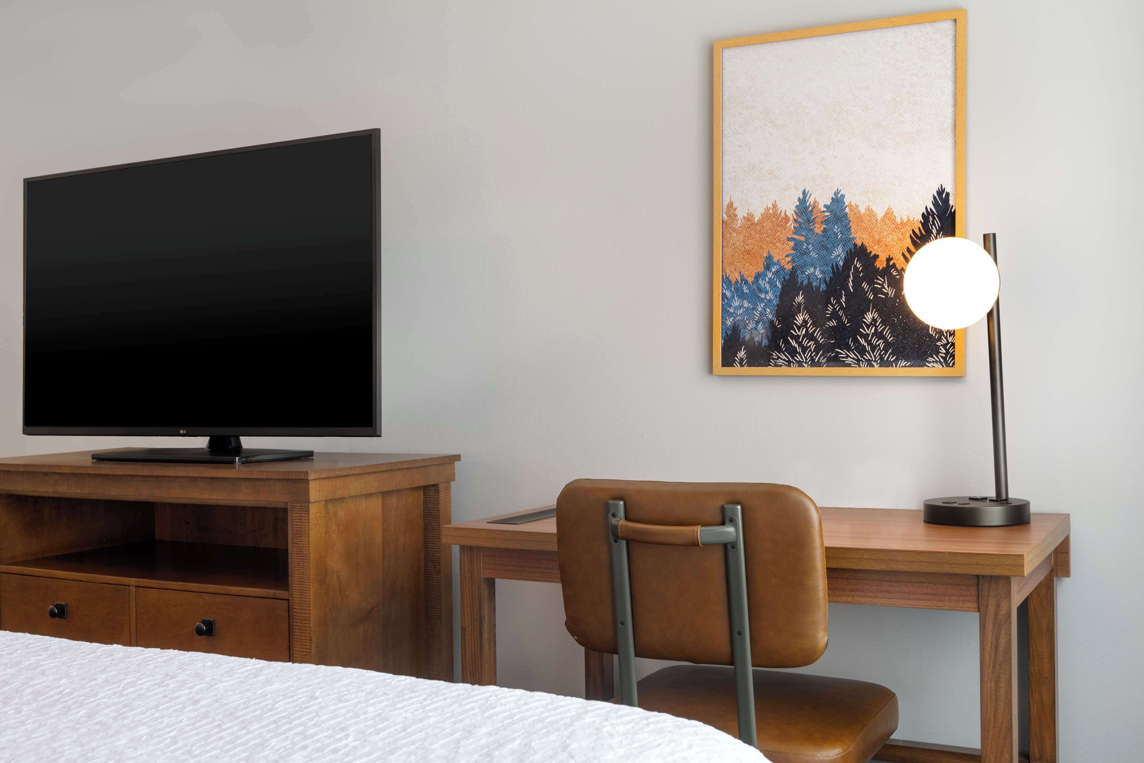 Quarto de hotel com cama, televisão em cómoda de madeira, escrivaninha com cadeira e quadro na parede.