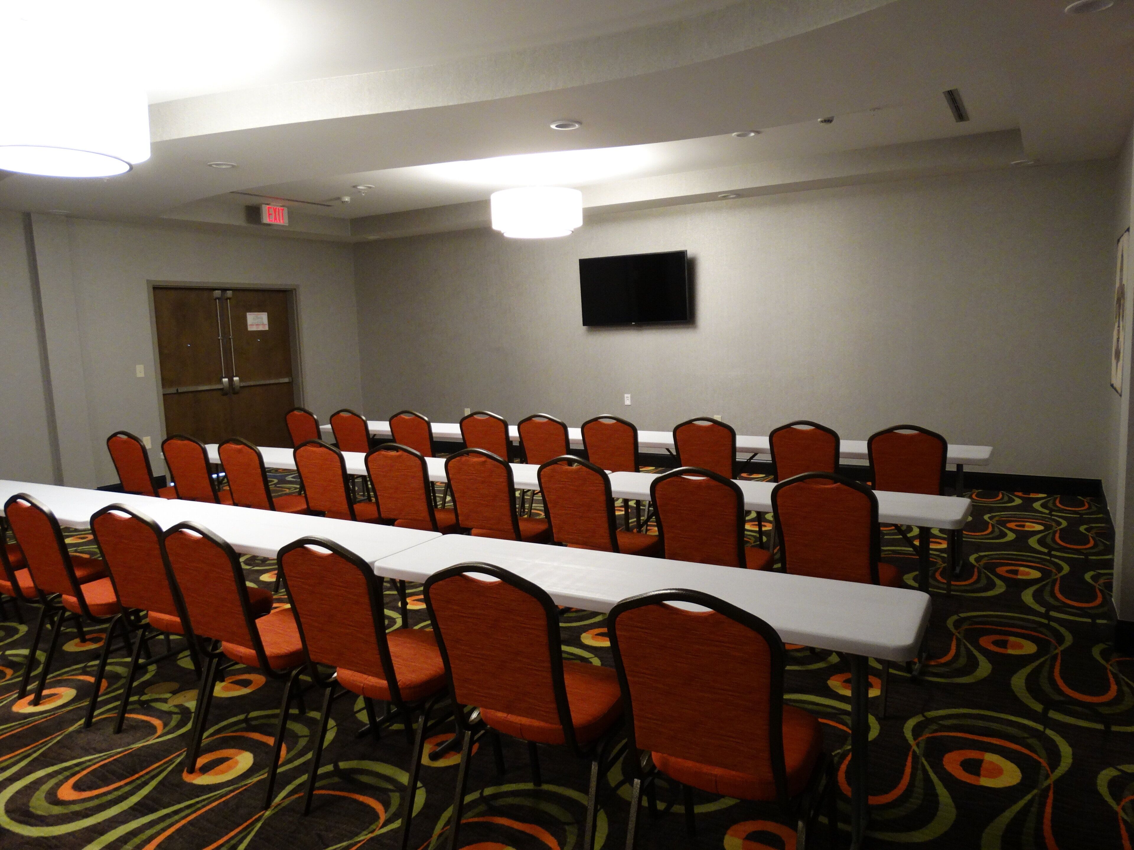 A conference room with a long table and rows of chairs, a door, and a flat-screen TV on the wall.