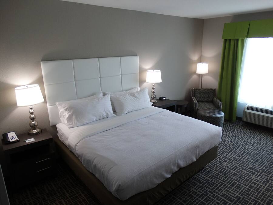 Quarto de hotel com uma cama grande, duas mesas de cabeceira, uma cadeira, um abajur e uma janela com cortinas.