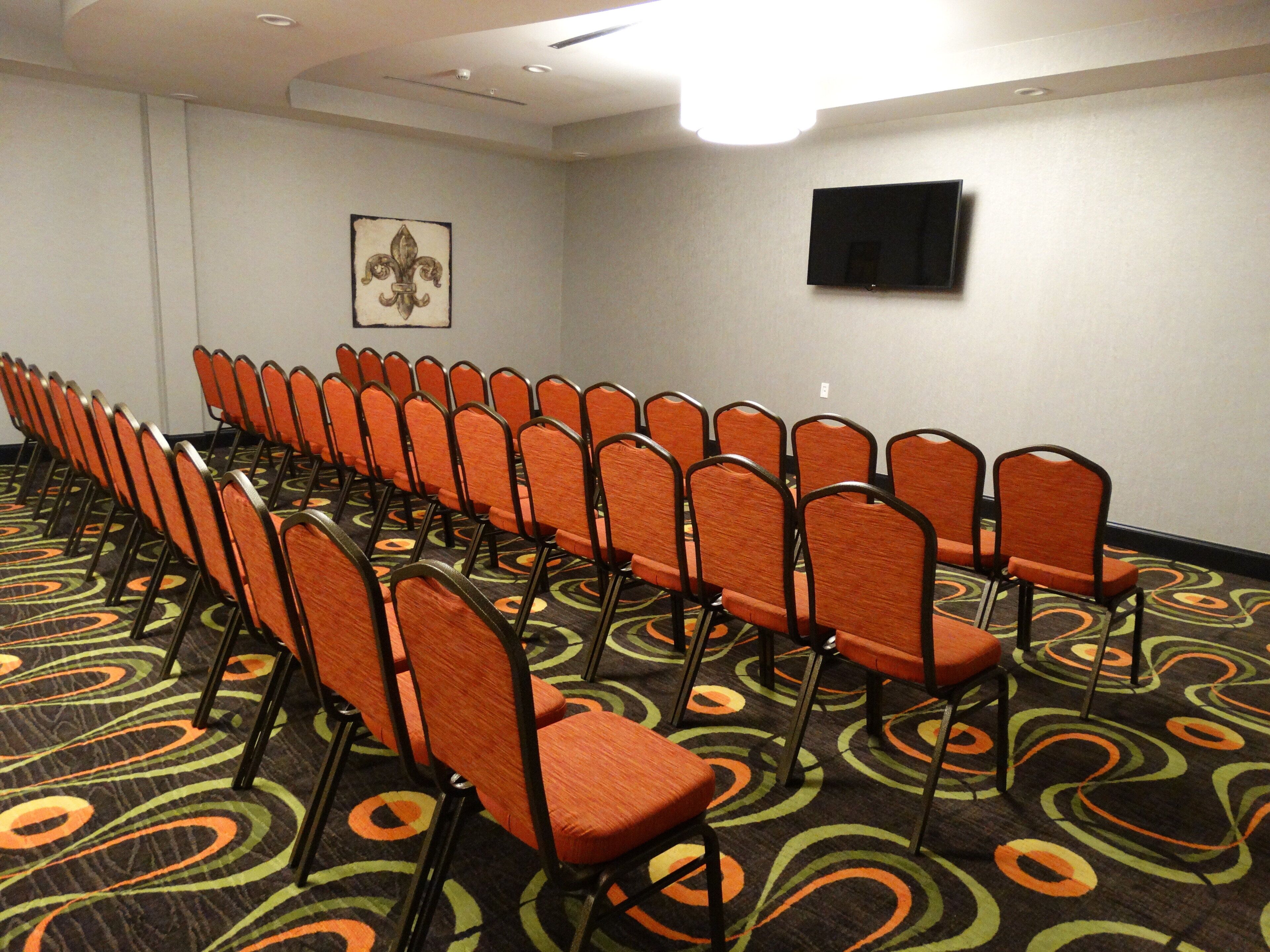 Une salle de conférence avec des rangées de chaises orange, un tapis orné de motifs circulaires, un téléviseur fixé au mur et un tableau encadré.
