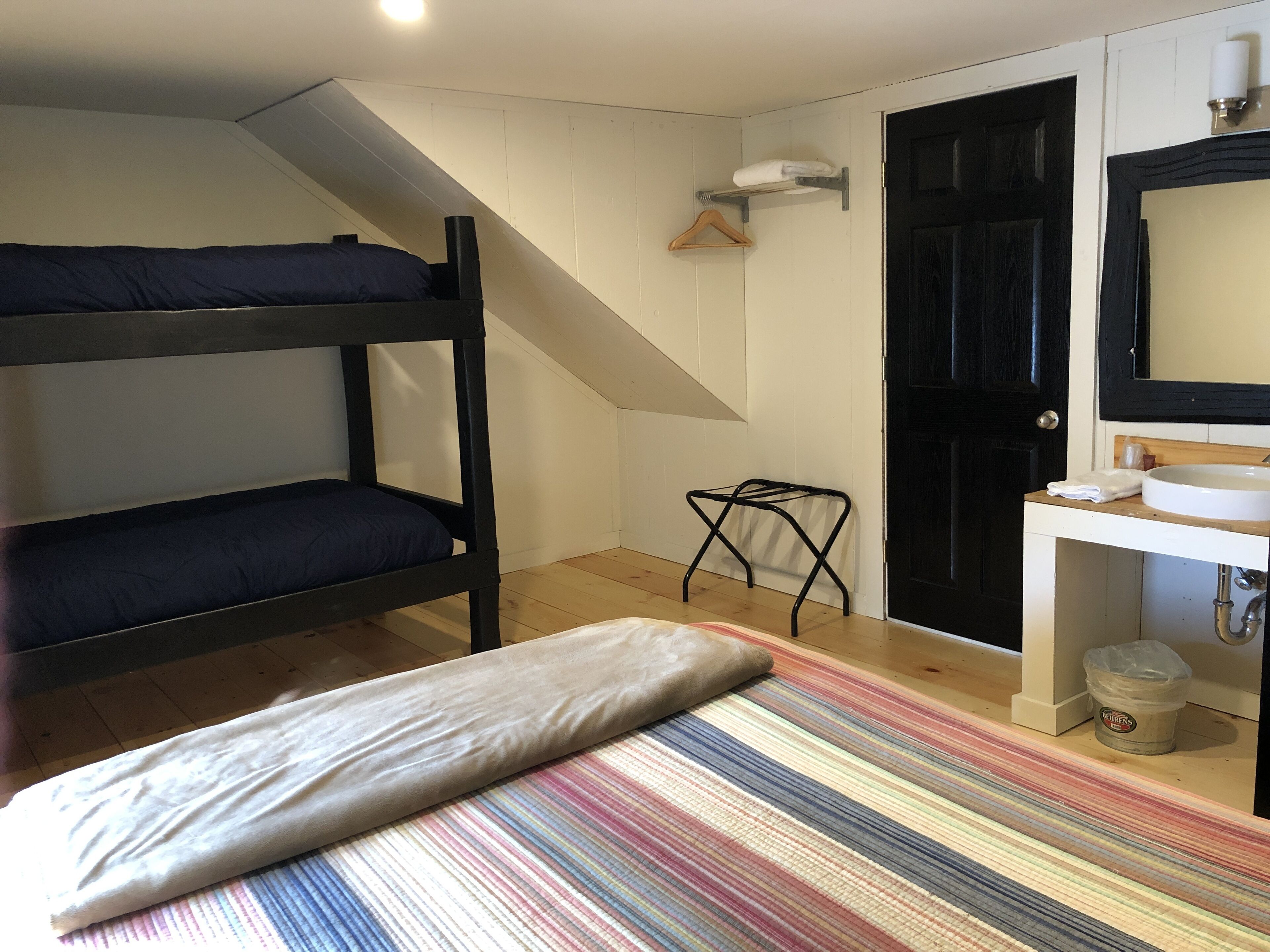A room with bunk beds, a small stool, a striped rug, a sink, and a door.