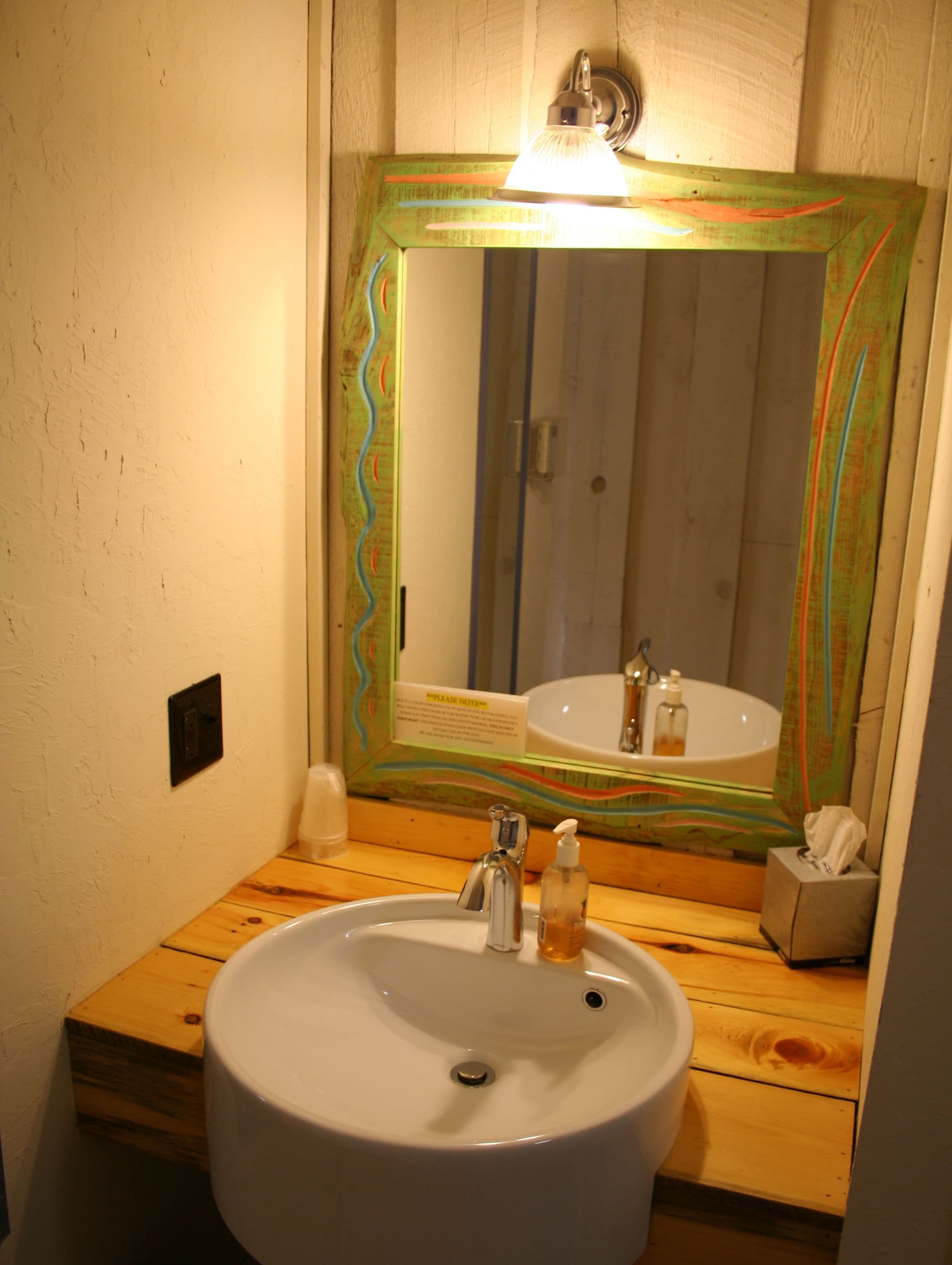 A bathroom with a wooden vanity, a white sink, a mirror with a colorful frame, and a "Please Note" sign.