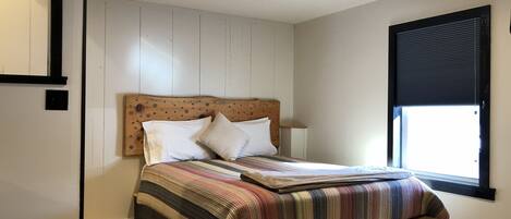 A bedroom with a wooden headboard, a striped bedspread, and a window with blinds.