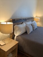 Un dormitorio con cabecera de madera, cama con ropa de cama blanca y gris, dos lámparas de noche y una mesita de noche con una lámpara y una bandeja.