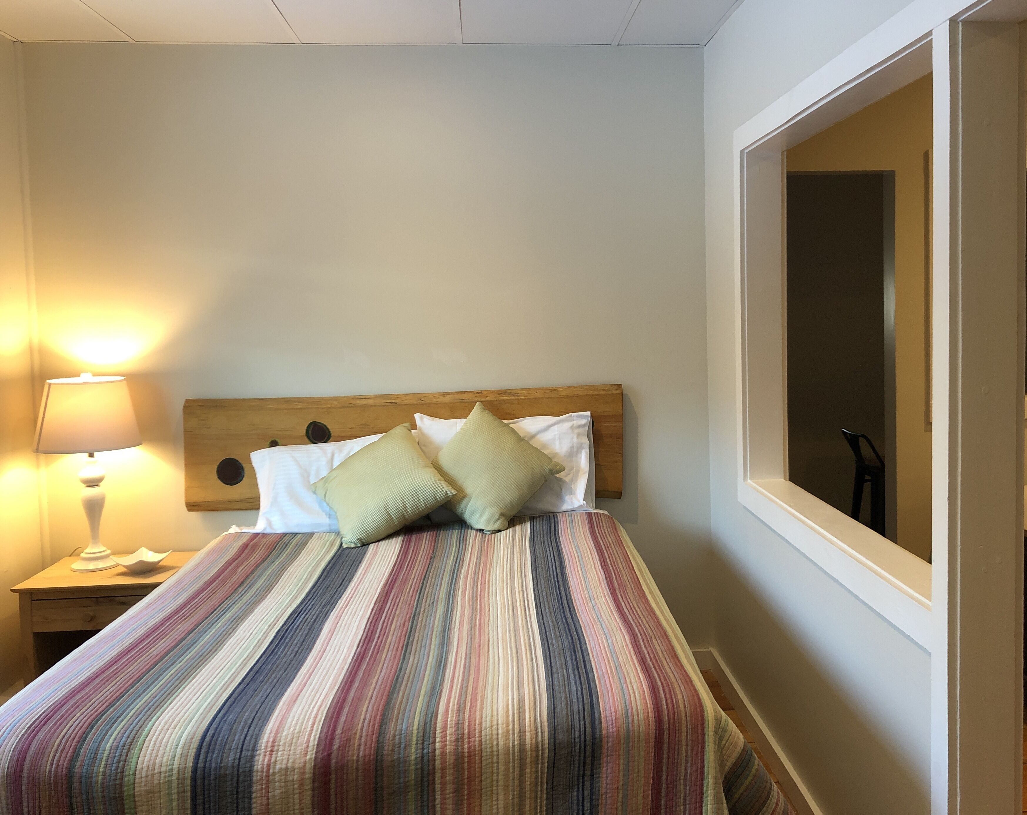 Una cama bien tendida con una colcha a rayas, dos almohadas, un cabecero de madera, una lámpara de noche y una ventana con vistas al exterior.