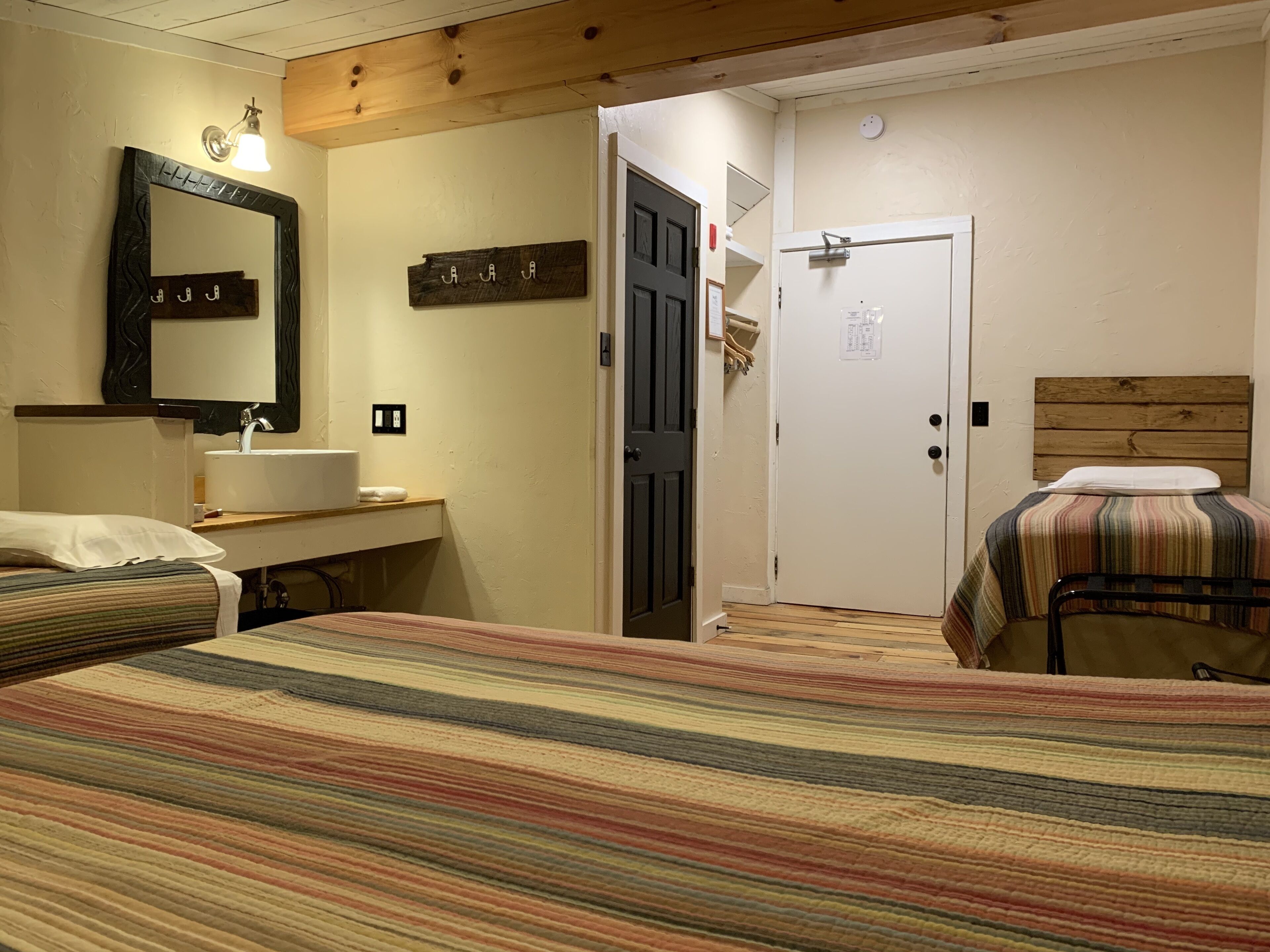 A room with a bed, a bedside table, a mirror, a door, and a wall-mounted light fixture.