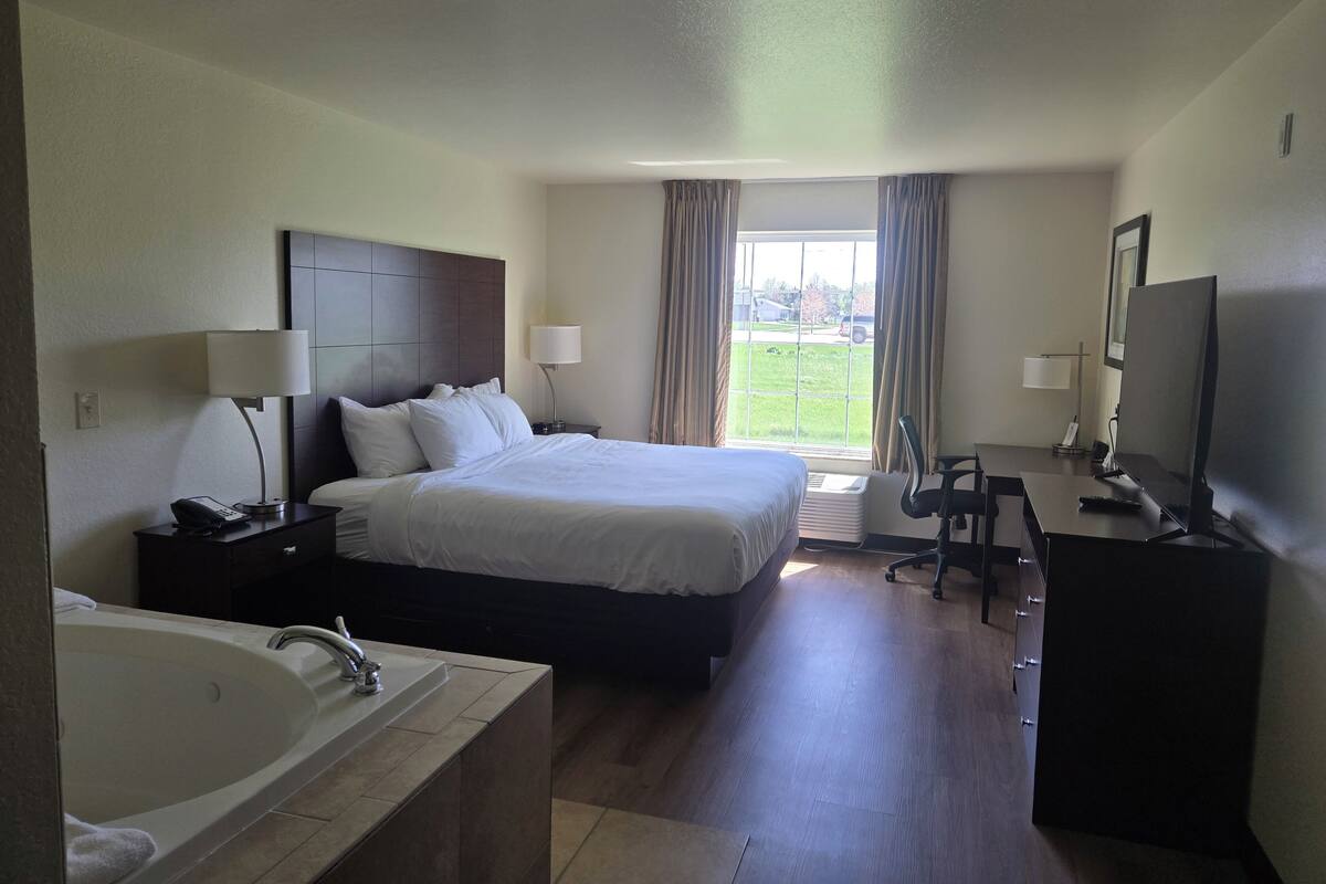 A hotel room with a large bed, a desk, a TV, a bathroom with a sink and bathtub, and a window with curtains.