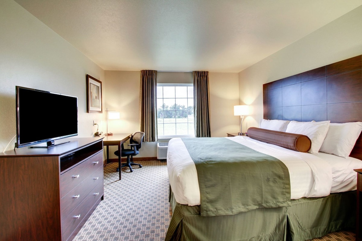 A hotel room with a bed, a desk with a TV, a chair, a window with curtains, and an air conditioning unit.