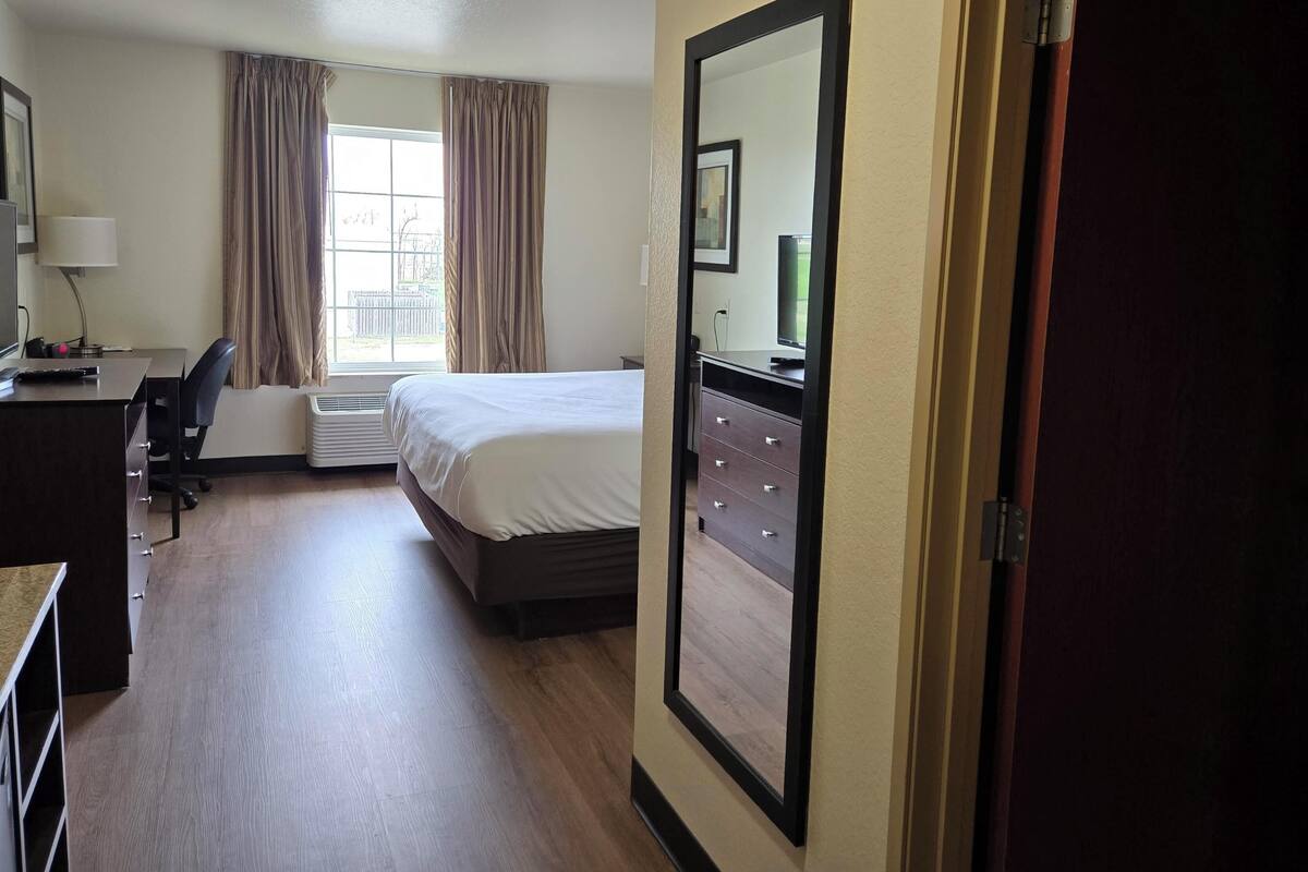 A hotel room with a bed, a desk with a chair, a dresser, a window with curtains, and a large mirror on the wall.