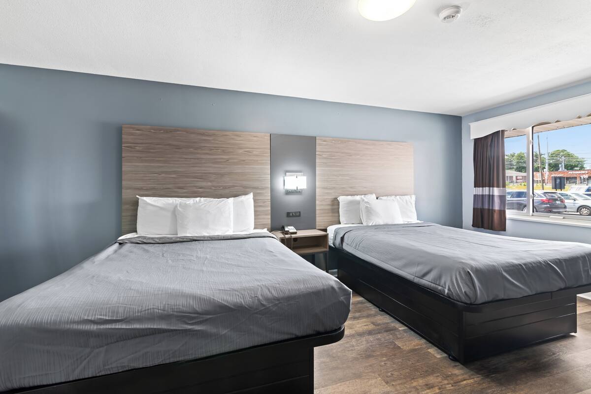 A hotel room with two beds, a nightstand, a wall-mounted light, and a window with a view of the street.