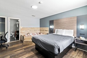 A modern hotel room with a large bed, a desk with a telephone, a chair, a bathtub, and a toilet visible through an open door.