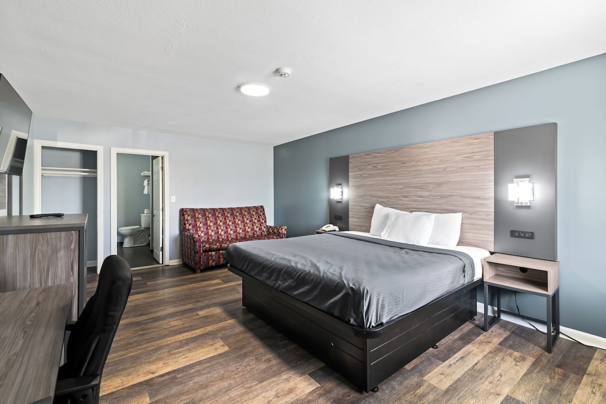 A modern hotel room with a large bed, a desk, a chair, a red patterned sofa, and a bathroom visible through an open door.
