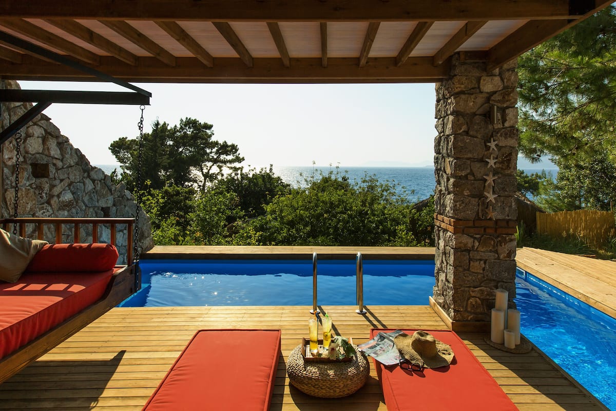 Una terraza de madera con piscina, cojines rojos y un muro de piedra.
