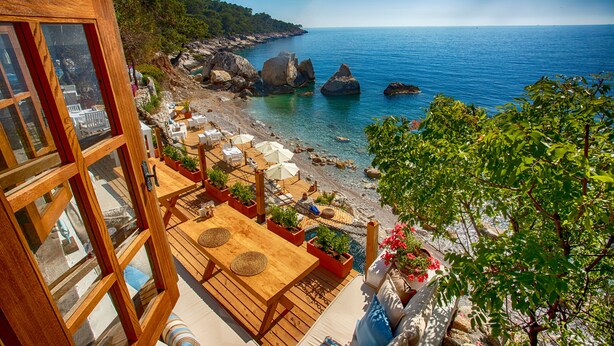 Eine Holzterrasse mit Sitzgelegenheiten, mit Blick auf einen Strand mit klarem, blauem Wasser und Felsen.